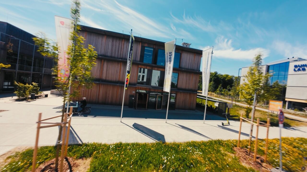 Das Gebäude der Energieagentur Regensburg mit Holzfassade und Fahnenmasten, aufgenommen aus der FPV-Drohne im Anflug