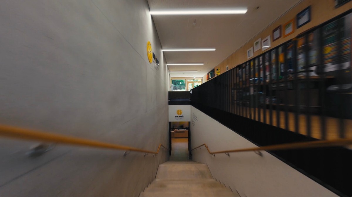 Flug über eine Treppe im Inneren, unten das Schild der Ausstellung Um:Welt, rechts eine Wand mit gerahmten Bildern