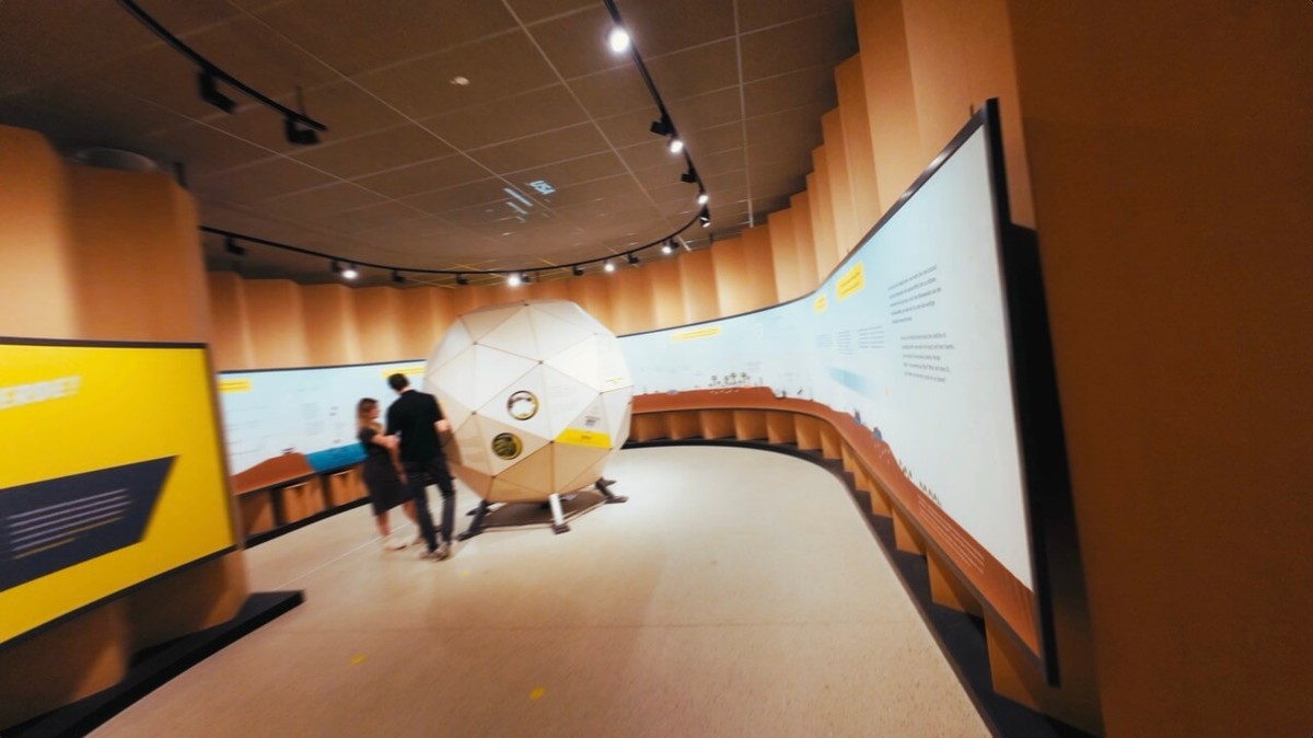 Ausstellungsraum Um:Welt mit einer großen weißen Kugel in der Mitte, zwei Besuchende davor, ringsum eine geschwungene Infowand