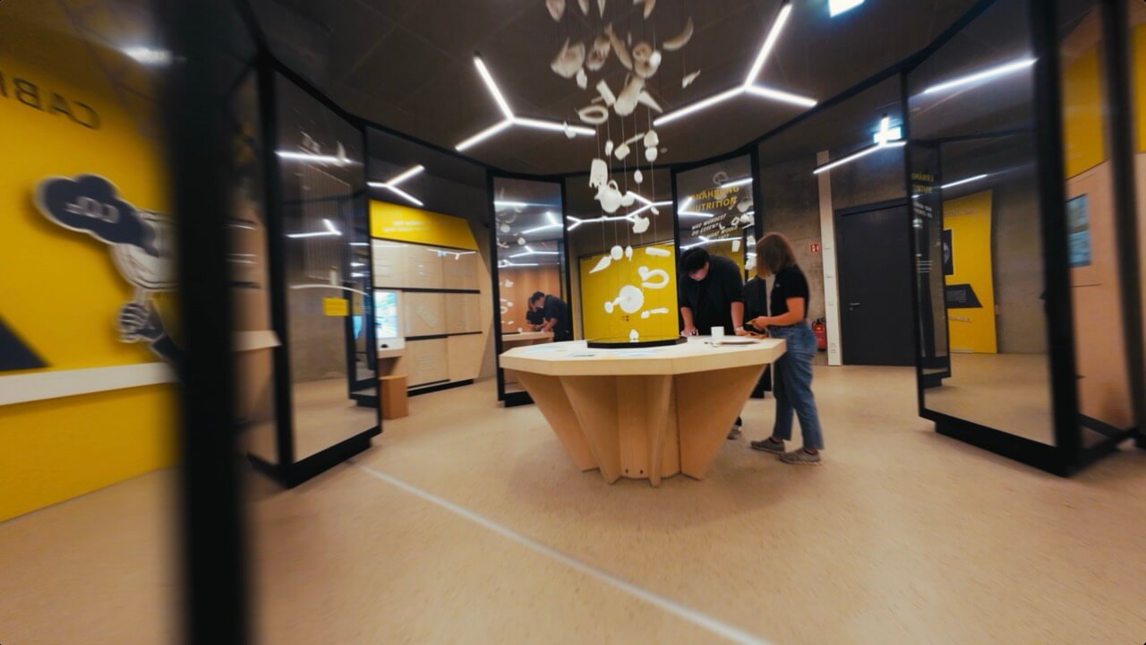 Ausstellungsbereich mit gelben Vitrinen und einem Mitmachtisch, an dem zwei Personen stehen, darüber ein Mobile aus weißen Objekten