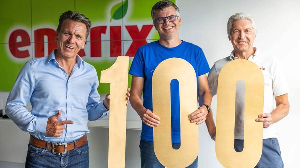 Drei Männer halten große Ziffern, die zusammen die Zahl 100 ergeben, dahinter das grüne enerix-Logo an der Wand