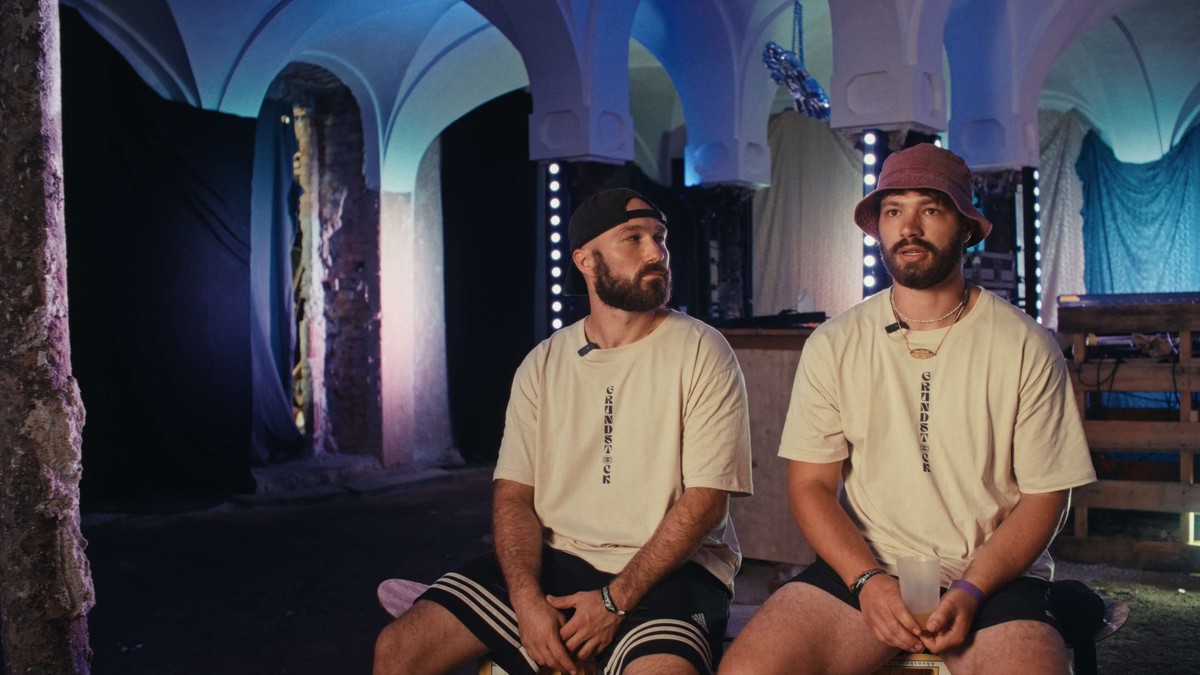 Zwei Veranstalter des Grundstock Festivals im Interview, in Shirts mit dem Festivalschriftzug, hinter ihnen ein Gewölbekeller in blauem Licht
