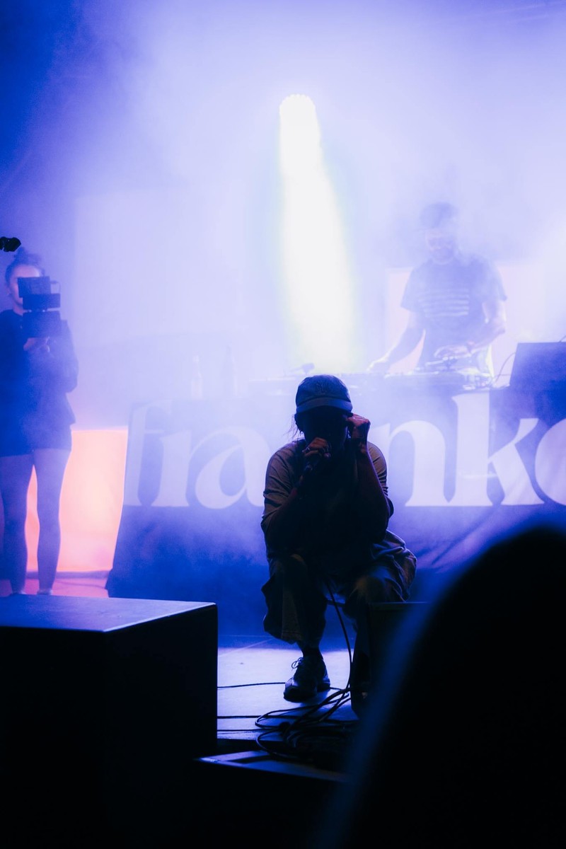 Rapper in der Hocke am Bühnenrand im blauen Scheinwerferlicht, im Hintergrund der DJ und eine Kamerafrau