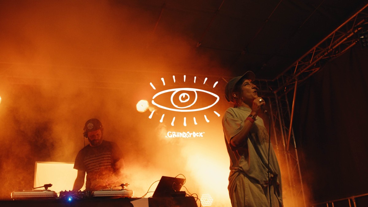 DJ und Sängerin auf der Bühne im orangen Nebel, dazwischen das gezeichnete Auge als Logo des Grundstock Festivals